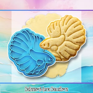 May include: A bright blue plastic betta fish cookie cutter positioned next to a baked beige cookie shaped like a betta fish with detailed fin and body impressions. The text CrimsonMane Creations is printed in black at the bottom of the image.