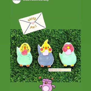May include: Three decorated sugar cookies shaped like parakeets, each holding a heart. The cookies are on a green grass background. An envelope with the words "Happy Mail" is above the cookies. A pink cartoon bird with hearts around its head is at the bottom of the image. The text "@crimson_mane_creations" is below the cookies.