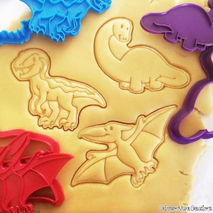 Könnte beinhalten: Nahaufnahme von Keksteig mit Dinosaurier-Ausstechern und bunten Ausstechformen aus Kunststoff. Die Ausstecher umfassen einen Tyrannosaurus Rex, einen Brontosaurus und einen Pterodactylus. Die Ausstecher sind blau, rot und lila.