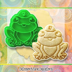 May include: A green frog-shaped cookie cutter next to a frog-shaped cookie. The cookie cutter is made of green plastic. The cookie is light brown and has a detailed frog design. The text "Crimson Mane Creations" is at the bottom.