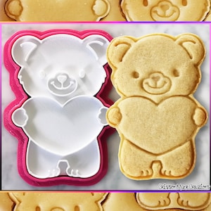 Può includere: Un tagliapasta di plastica rosa a forma di orso che tiene un cuore. Accanto al tagliapasta c'è un biscotto cotto a forma di orso che tiene un cuore.