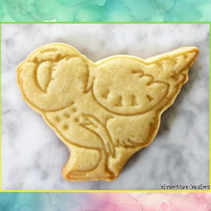 May include: A light-colored, heart-shaped cookie with an embossed design of a bird. The cookie has a detailed outline and texture, with the words "Crimson Mare Creations" in the bottom right corner. The cookie is on a white surface.