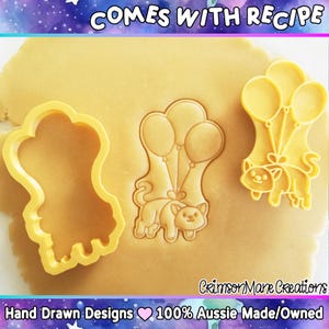 Pu&ograve; includere: Tre formine per biscotti in plastica gialla: un gatto con palloncini, un contorno di gatto e un gatto con palloncini. Le formine sono su una superficie marrone chiaro. Il testo include "COMES WITH RECIPE", "Crimson Mane Creations" e "Hand Drawn Designs 100% Aussie Made/Owned".