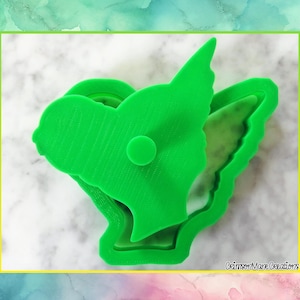May include: A bright green cookie cutter in the shape of a fairy or angel with wings. The cutter has a round button on the top and is made of plastic. The text "Crimson Mane Creations" is in the bottom right corner.