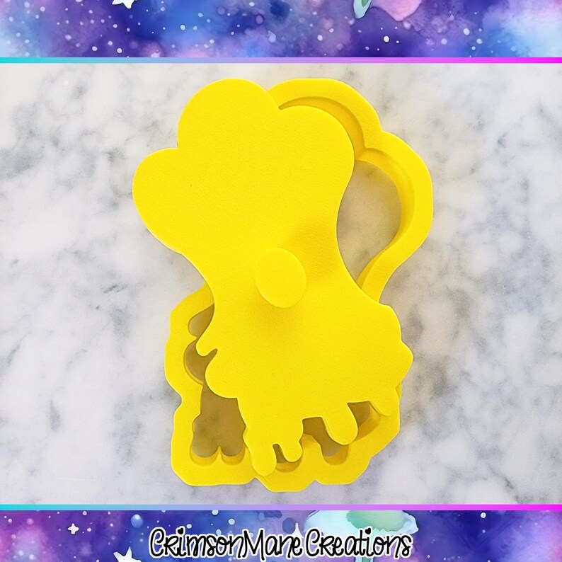 Pu&ograve; includere: Tagliabiscotti giallo brillante a forma di personaggio dei cartoni animati. Il tagliabiscotti ha una parte superiore arrotondata con un piccolo cerchio e un bordo inferiore ondulato. Il testo "Crimson Mane Creations" &egrave; in basso.