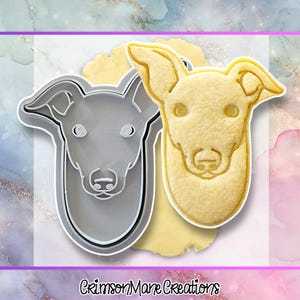 May include: A grey plastic dog-shaped cookie cutter next to a matching light beige sugar cookie. The design features the head of a greyhound with pointed ears and a snout. The text "CrimsonMane Creations" is printed at the bottom of the image.