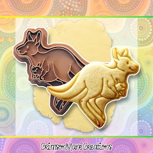 May include: A brown kangaroo cookie cutter next to a kangaroo-shaped cookie. The cookie cutter is made of plastic and has a detailed design of a kangaroo with a joey in its pouch. The cookie is light brown and has the same kangaroo shape.