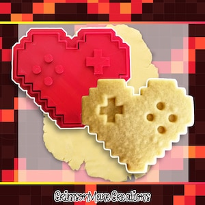 May include: A red plastic heart-shaped cookie cutter with a pixelated design featuring a D-pad and four circular buttons, alongside a matching baked cookie made from dough, with the text CrimsonManeCreations visible at the bottom of the image.