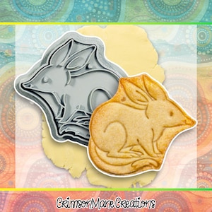 Könnte beinhalten: Ein grauer Metall-Ausstecher in Form eines Bilbys, neben einem gebackenen Keks in Bilby-Form. Der Keks ist goldbraun mit detaillierten Merkmalen. Der Hintergrund ist ein farbenfrohes, gemustertes Design mit dem Text "Crimson Mane Creations".