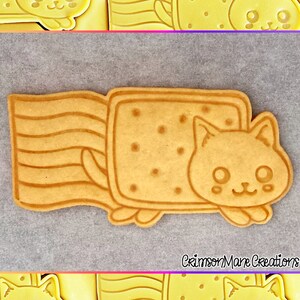May include: A tan-colored cookie cutter in the shape of a cat emerging from a rectangular cookie. The rectangular cookie has small dot indentations. The cat has a smiling face and small paws. The cookie cutter is on a gray surface.
