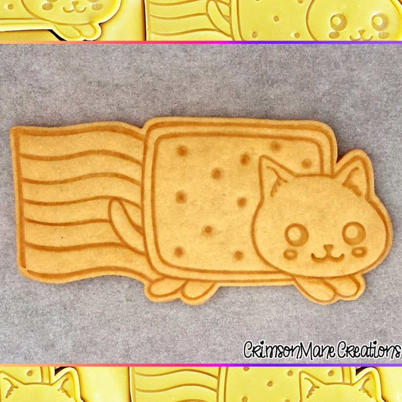 May include: A tan-colored cookie cutter in the shape of a cat emerging from a rectangular cookie. The rectangular cookie has small dot indentations. The cat has a smiling face and small paws. The cookie cutter is on a gray surface.