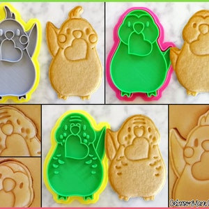 May include: A set of four cookie cutters in the shape of a parakeet holding a heart. Two cutters are green, one is gray, and one is brown. The cutters are on a white surface.
