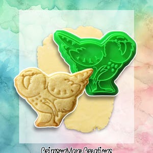 May include: A green plastic cookie cutter in the shape of a bird sits next to a bird-shaped cookie. The cookie is light brown and has detailed markings. The cookie cutter is on a light yellow surface, with a colorful background.