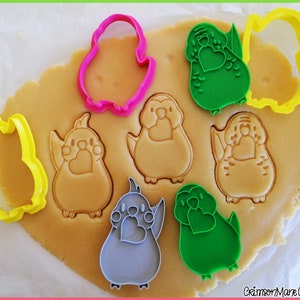 May include: A set of cookie cutters in the shape of a bird holding a heart. The cutters are pink, green, yellow, gray, and blue. The cutters are on a piece of cookie dough.
