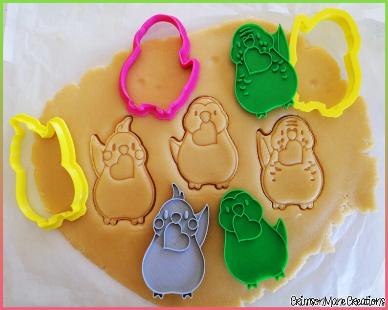 May include: A set of cookie cutters in the shape of a bird holding a heart. The cutters are pink, green, yellow, gray, and blue. The cutters are on a piece of cookie dough.