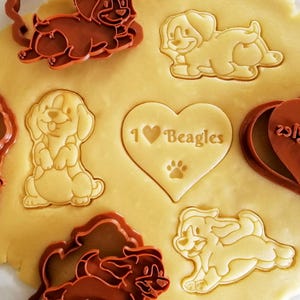 Könnte beinhalten: Nahaufnahme von Keksteig mit mehreren hundeformigen Ausstechern und gestempelten Keksen. Ein Keks ist herzförmig mit dem Text "I ❤️ Beagles" und einem Pfotenabdruck. Die Ausstecher sind braun und die Kekse hellgelb.