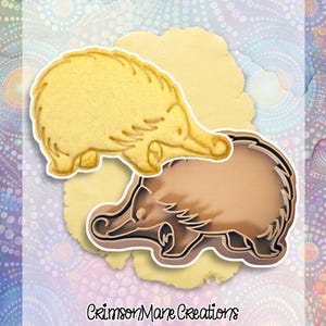 May include: An echidna-shaped cookie cutter and a baked cookie. The cookie cutter is brown with a white outline, and the cookie is golden yellow. The background includes a light yellow dough and a patterned design with the text "Crimson Mane Creations".