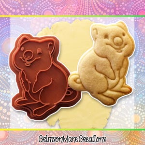 May include: A brown plastic cookie cutter and a baked cookie, both in the shape of a wombat. The cookie cutter has a detailed outline of the animal. The baked cookie is light brown and has a smiling face. The text "Crimson Mane Creations" is at the bottom of the image.