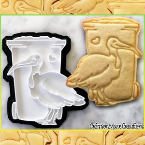 Peut inclure: Un emporte-pièce en plastique blanc en forme de poubelle avec un oiseau à l'intérieur. L'emporte-pièce est sur une surface blanche à côté d'un biscuit brun au sucre de la même forme. Le biscuit a un glaçage blanc en forme d'oiseau à l'intérieur de la poubelle.
