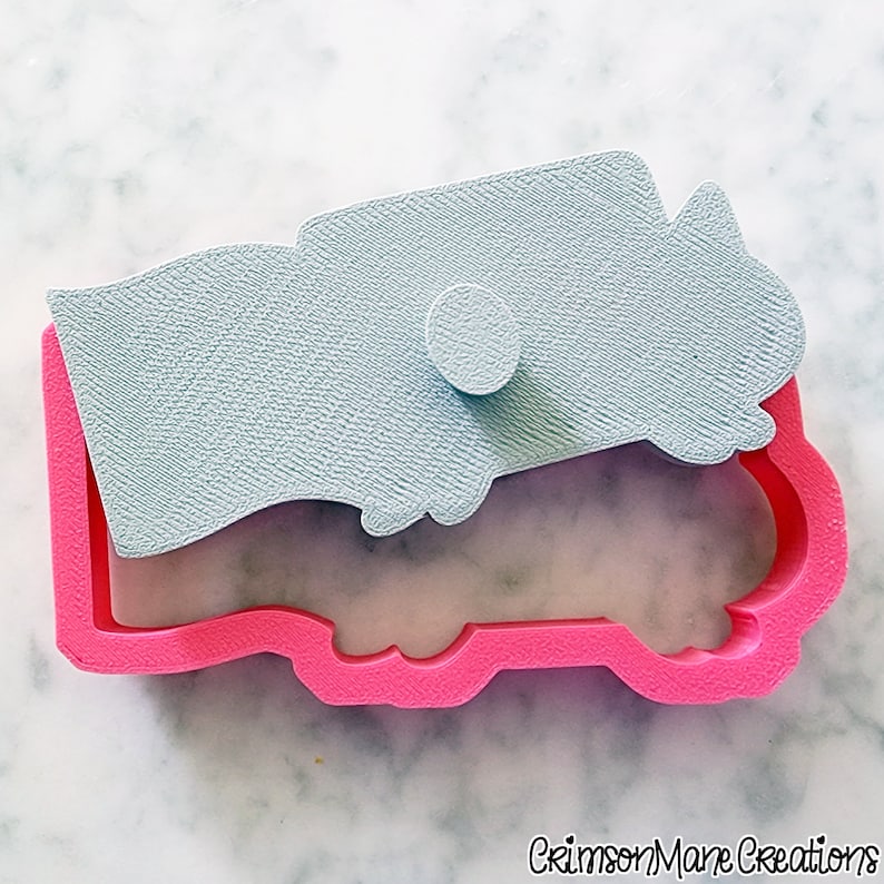 May include: A pink cookie cutter in the shape of a truck with a light blue textured stamp and a small round button. The cookie cutter is on a white marble surface. The text "Crimson Mane Creations" is in the bottom right corner.
