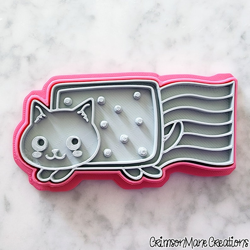 May include: A pink, gray, and white cat-shaped cookie cutter. The cat has a rectangular body with a rainbow tail. The cat's face has large eyes and a small nose. The cookie cutter is made of plastic and has a layered design.