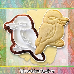 May include: A bird-shaped cookie cutter and a baked cookie. The cutter is white with a brown outline. The cookie is light brown and sits on a piece of dough. The text "Crimson Mane Creations" is at the bottom of the image.