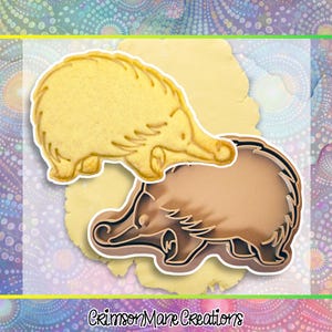 May include: An echidna-shaped cookie cutter and a biscuit. The cutter is brown, and the biscuit is yellow. The cutter is next to a biscuit cut from dough. The text "Crimson Mane Creations" is at the bottom of the image.