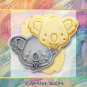 Könnte beinhalten: Ein grauer Ausstechform in Koala-Form und ein passender gebackener Keks in Koala-Form. Die Ausstechform zeigt den detaillierten Umriss eines Koala-Gesichts mit großen Ohren und einem Blatt im Mund. Der Text CrimsonMane Creations steht unten.