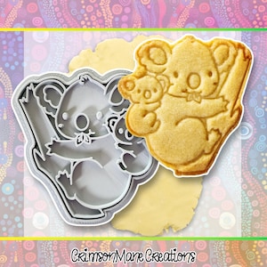 May include: A koala bear cookie cutter next to a baked koala bear cookie. The cookie cutter is gray and shaped like a koala bear holding a baby koala. The baked cookie is light brown and shows the koala bear holding a baby koala.