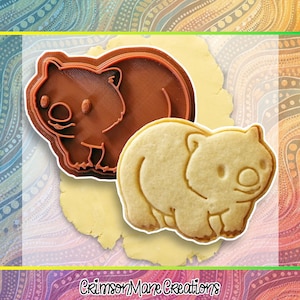 May include: A brown wombat cookie cutter and a wombat-shaped biscuit. The cutter is plastic and has a detailed outline. The biscuit is light brown and has a detailed imprint of the wombat. The image includes the text "Crimson Mane Creations".