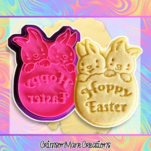 May include: A pink and purple Easter-themed cookie cutter with a bunny design and the words "Happy Easter" in reverse. Next to it is a baked cookie with the same design. The cookie cutter is made of plastic and the cookie is light yellow.