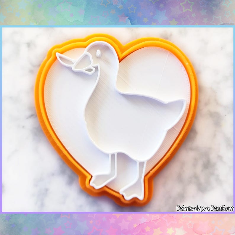 May include: A white duck-shaped cookie cutter inside an orange heart-shaped frame. The cookie cutter is a detailed outline of a duck with a beak, wings, and feet. The design is set against a white background.