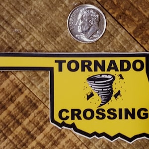 May include: Yellow sticker shaped like the state of Oklahoma with the words "Tornado Crossing" and a black and white illustration of a tornado with cows flying around it.