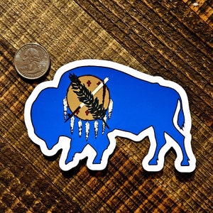 May include: A blue and white sticker of a buffalo with the state flag of Oklahoma on its back. The flag features a Native American shield with a peace pipe and an olive branch.