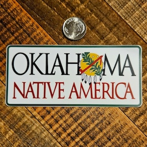 May include: A white sticker with green trim features the text "OKLAHOMA NATIVE AMERICA" in black and red lettering. A stylized Native American symbol with a yellow background, green leaves, and red and white feathers is centered above the text.