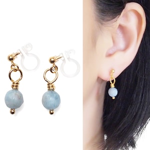 May include: A pair of clip-on earrings with a light blue gemstone dangle. The earrings are made with gold-toned metal and have a clear plastic clip.
