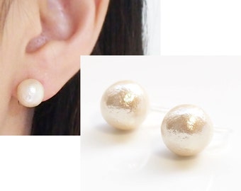 May include: A pair of round, cream-colored stud earrings with a textured, matte finish. The earrings feature a spherical design that mimics the appearance of a pearl. One earring is shown attached to an earlobe, while both are displayed together.