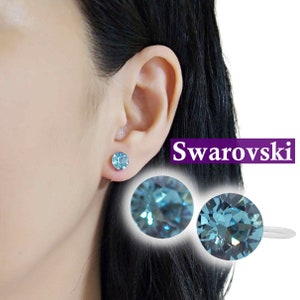 以下が含まれることがあります： シルバーのセッティングに青いクリスタルをあしらったスタッドピアスのペア。ピアスはモデルの耳に装着されています。テキスト「Swarovski」は紫色の文字で表示されています。