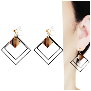 May include: Geometric earrings featuring a gold-colored square and a black square outline. The earrings have a gold-colored stud and a clear clip-on backing. The design is modern and minimalist.
