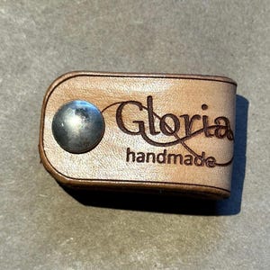May include: A small, light brown leather key holder with a silver metal snap. The word "Gloria" is engraved in a cursive font, with "handmade" below it. The leather has a natural, textured appearance.