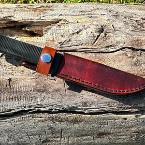 May include: A brown leather knife sheath with a stitched edge. The sheath is on a wooden surface.