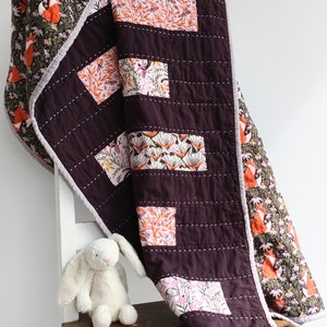 May include: A burgundy quilt with a white and pink floral pattern and a brown and green fox print. The quilt is folded over a wooden chair with a white bunny plush toy.