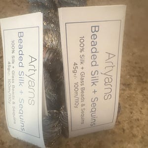May include: Two skeins of Artyarns Beaded Silk + Sequins yarn. The yarn is a dark gray color with gold sequins and beads. The labels read "100% Silk + Glass Beads & Sequins" and "45g+/- 100m/110y".