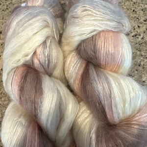 May include: Close-up of two skeins of yarn in shades of cream, pink, and brown. The yarn appears soft and fluffy, with a slight sheen. The colors blend together in a gradient effect, creating a visually appealing texture. Ideal for knitting or crochet projects.