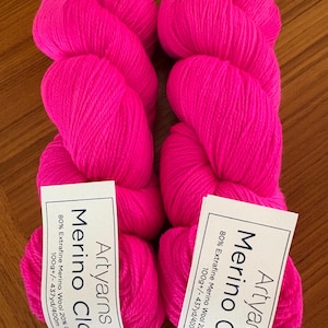May include: Two skeins of bright pink Merino Cloud yarn by Artyarns. Each skein has a label with the brand name and fiber content: 80% Extrafine Merino Wool, 20% Cashmere, 100g +/- 437yd/400m. The yarn is displayed on a wooden surface.