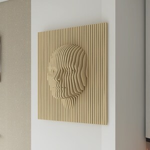 Könnte beinhalten: Eine Wandskulptur aus Holz, die ein Gesicht darstellt, aus dünnen Holzstreifen gefertigt, mit einem geschichteten Effekt. Die Skulptur ist an einer weißen Wand in einem modernen Interieur angebracht.