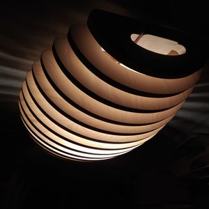 May include: A modern, wooden pendant light fixture. The light is composed of layered, curved wooden slats, creating a spiral design. The light emits a warm glow, contrasting with the dark background. The fixture has a circular handle at the top.