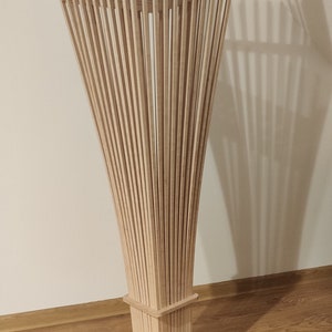 May include: A modern wooden floor lamp with a unique design. The lamp features numerous vertical wooden slats that flare outwards from a square base, creating a sculptural, artistic form. The lamp is made of light-colored wood.