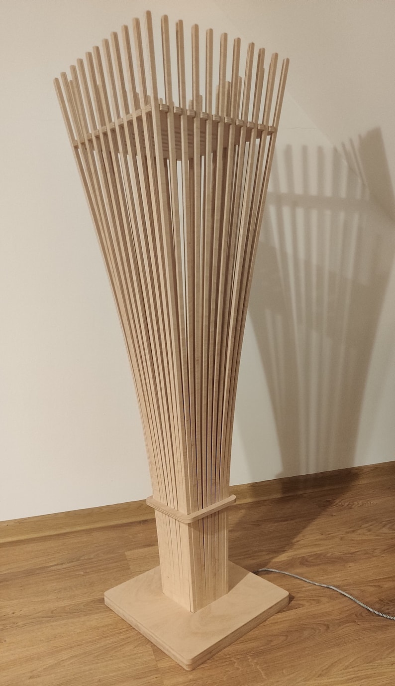 May include: A modern wooden floor lamp with a unique design. The lamp features numerous vertical wooden slats that flare outwards from a square base, creating a sculptural, artistic form. The lamp is made of light-colored wood.