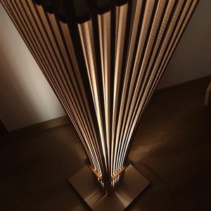 May include: A modern, geometric floor lamp with a square base and a fan-like shade. The lamp is made of light-colored wood and has a warm, inviting glow. The design features vertical wooden slats that create a unique lighting effect.